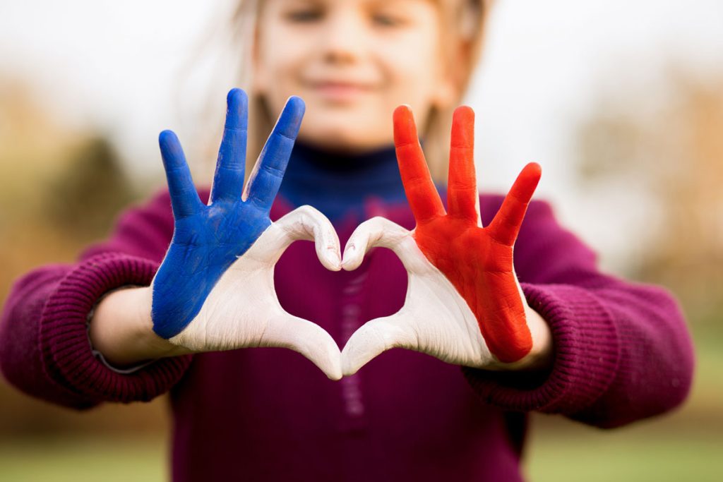 Little girl smiling, making a heart with her hands. Her hands are painted with the French flag.