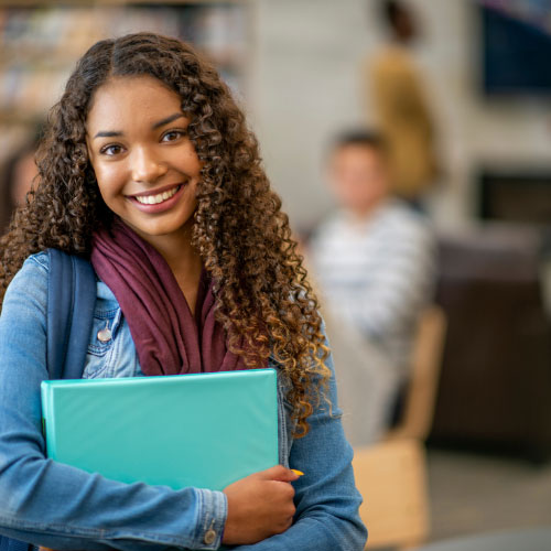 A teenage girl in a jean jacket facing the camera and smiling. She is holding some books. She aced her exam thanks to FLAM San Diego.