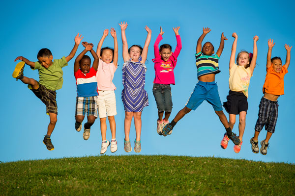 Colorful and fun image of several young kids jumping in the air on a green hill with their arms up.